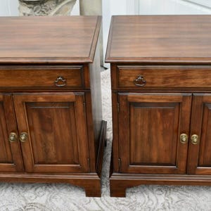 May include: Two matching wooden nightstands with a rich, dark brown finish. Each nightstand features a drawer with a decorative pull and two cabinet doors with brass-colored hardware. The nightstands are rectangular with a slightly overhanging top.