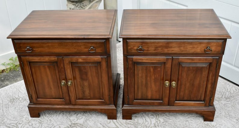 May include: Two matching wooden nightstands with a rich, dark brown finish. Each nightstand features a drawer with a decorative pull and two cabinet doors with brass-colored hardware. The nightstands are rectangular with a slightly overhanging top.