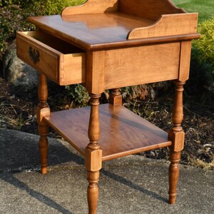 May include: A light brown wooden side table with a drawer and a lower shelf. The drawer is open, revealing the dovetail construction. The table has turned legs and a decorative top edge. The table is approximately 30 inches tall.