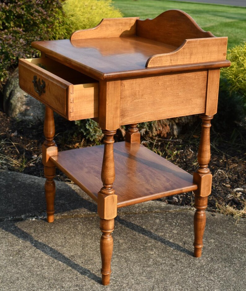 May include: A light brown wooden side table with a drawer and a lower shelf. The drawer is open, revealing the dovetail construction. The table has turned legs and a decorative top edge. The table is approximately 30 inches tall.
