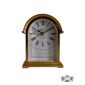 May include: A gold-toned mantel clock with an arched top and a white face featuring black Roman numerals. The clock displays the brand name Bulova and the word Quartz on the dial, with black hour, minute, and second hands. The word Taiwan is printed at the bottom of the clock face.