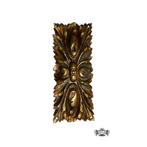 May include: Ornate, rectangular, gold-toned decorative architectural element with a symmetrical leaf design. The piece has a weathered appearance, with darker accents in the recessed areas. It features three small holes for mounting.
