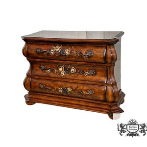 May include: A three-drawer wooden chest with a dark brown finish. The drawers feature floral designs and ornate handles. The chest has a curved design and sits on short legs. The top is a darker color than the rest of the chest.