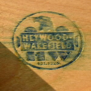 May include: Close-up of a wooden surface with a circular blue stamp. The stamp reads "HEYWOOD-WAKEFIELD" with an eagle emblem and "EST. 1826" below. The wood has a light brown color.