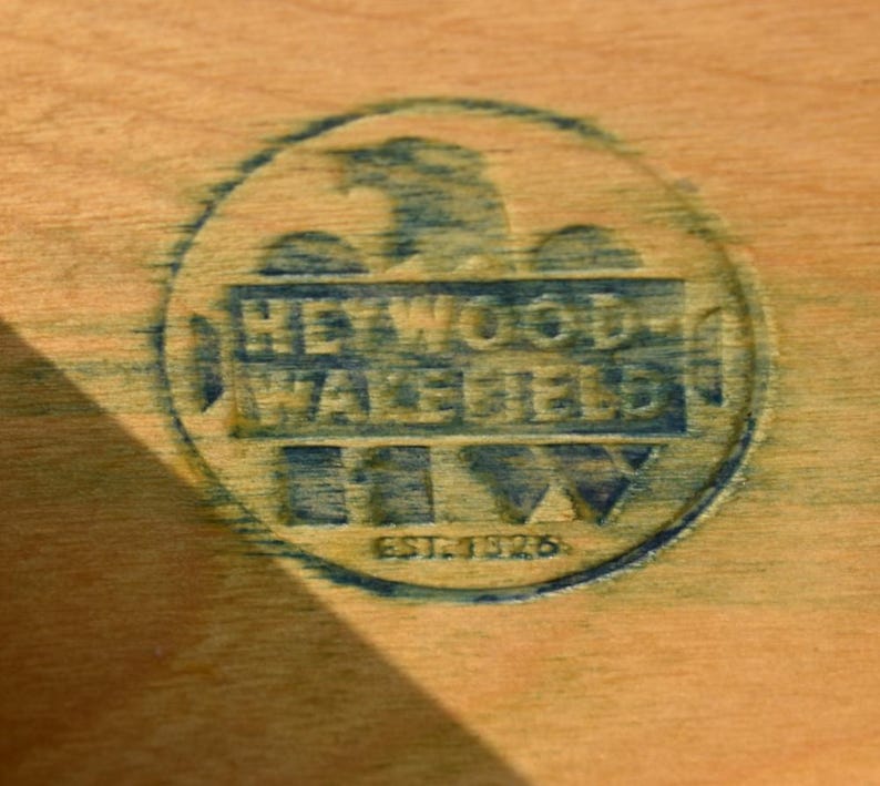 May include: Close-up of a wooden surface with a circular blue stamp. The stamp reads "HEYWOOD-WAKEFIELD" with an eagle emblem and "EST. 1826" below. The wood has a light brown color.