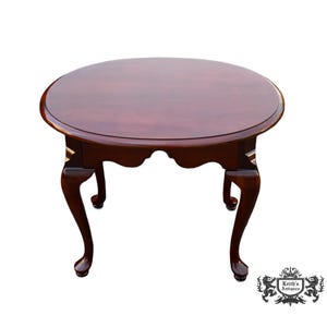 May include: A round, dark cherry wood coffee table with a glossy finish. The table features a scalloped apron and four curved legs. The legs have a slight cabriole shape. The table is approximately 30 inches in diameter.
