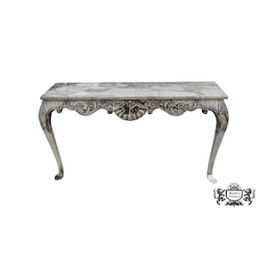 May include: A marble-topped antique table with ornate, carved detailing. The table features a gray and white marble surface and curved legs. The apron of the table is intricately carved with floral and shell motifs. The table is a classic piece of furniture.