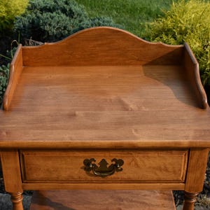 May include: A wooden side table with a warm, medium-brown finish. It features a single drawer with a decorative brass handle and a scalloped back. The table has a rectangular top and stands on four legs.