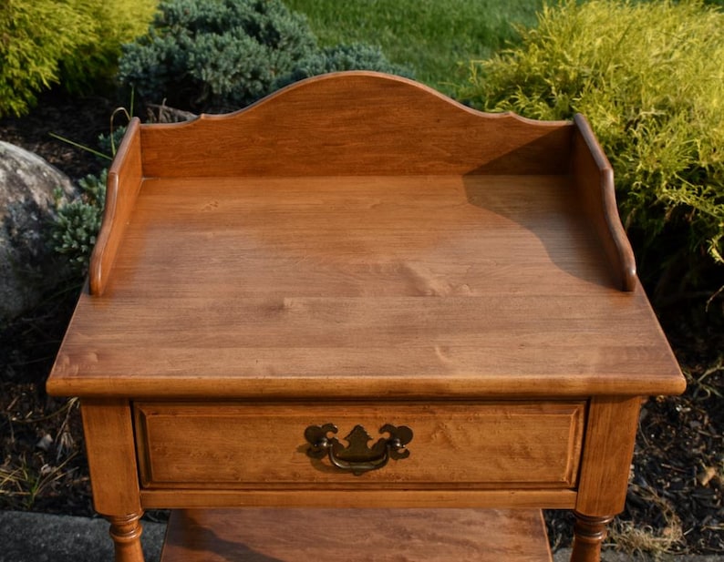 May include: A wooden side table with a warm, medium-brown finish. It features a single drawer with a decorative brass handle and a scalloped back. The table has a rectangular top and stands on four legs.
