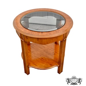May include: A round, antique side table with a glass top and a lower shelf. The table is made of light brown wood with a glossy finish. The table is approximately 24 inches in diameter and 28 inches tall.