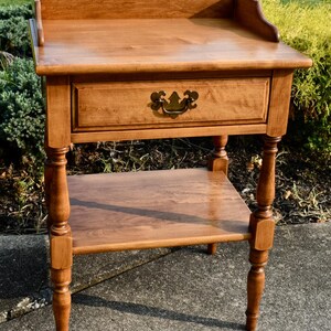 May include: A wooden side table with a drawer and a lower shelf. The table is made of light brown wood and has a decorative handle on the drawer. The table has a curved back and turned legs. The table measures approximately 28 inches high.
