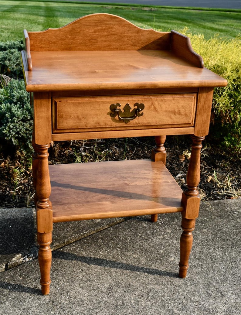 May include: A wooden side table with a drawer and a lower shelf. The table is made of light brown wood and has a decorative handle on the drawer. The table has a curved back and turned legs. The table measures approximately 28 inches high.