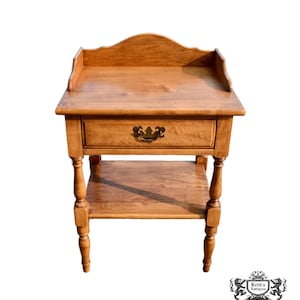 May include: A light brown wooden side table with a drawer and a lower shelf. The table has a curved top and ornate drawer pull. The legs are turned and the table is approximately 28 inches tall.