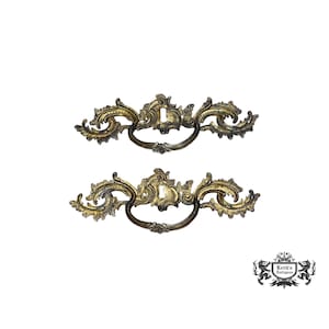 May include: Two ornate gold-toned metal drawer pulls with a decorative scroll design.
