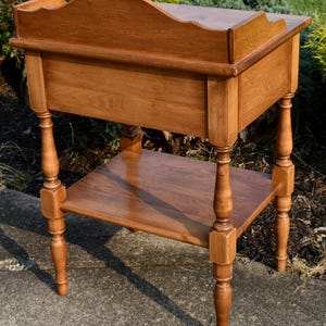 May include: A wooden side table with a warm, medium-brown finish. The table features a scalloped top, a lower shelf, and turned legs. The design is classic and elegant, suitable for various decor styles. The table measures approximately 30 inches tall.
