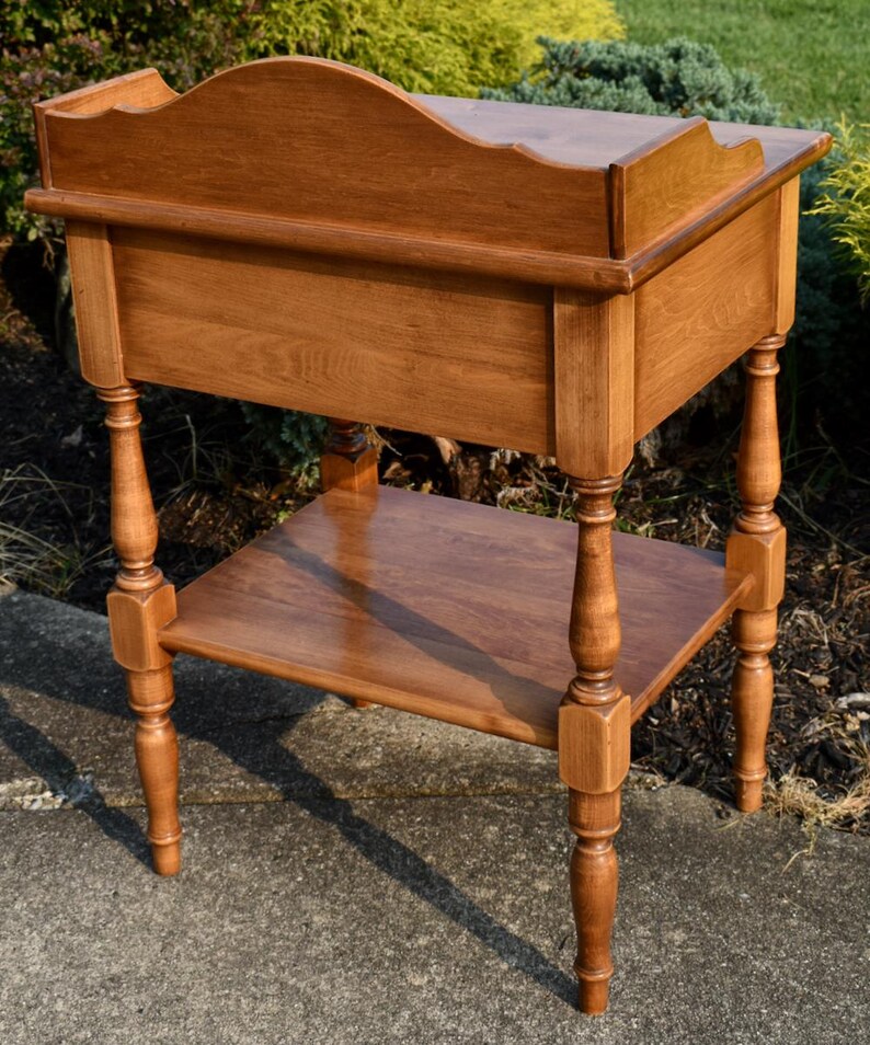 May include: A wooden side table with a warm, medium-brown finish. The table features a scalloped top, a lower shelf, and turned legs. The design is classic and elegant, suitable for various decor styles. The table measures approximately 30 inches tall.