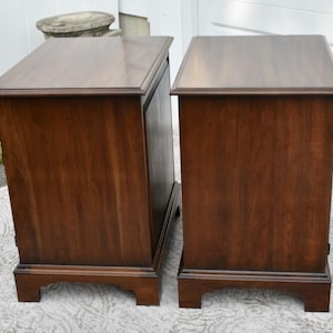 May include: Two matching wooden nightstands with a rich, dark brown finish. Each nightstand features a rectangular top, a single drawer, and a decorative base. The nightstands are placed side by side, showcasing their similar design and construction.