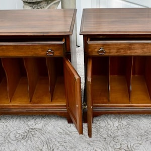 May include: Two matching wooden nightstands with drawers and cabinet doors. The nightstands are a warm brown color with decorative hardware. The cabinet doors are open, revealing interior shelving.