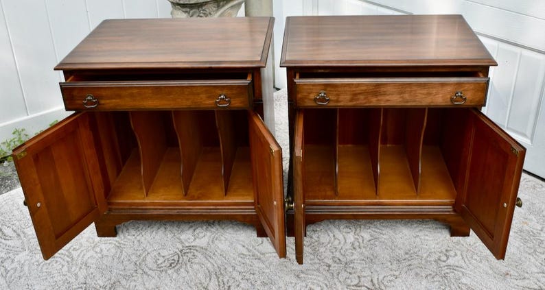 May include: Two matching wooden nightstands with drawers and cabinet doors. The nightstands are a warm brown color with decorative hardware. The cabinet doors are open, revealing interior shelving.