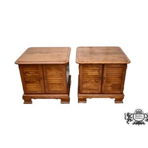 May include: Two matching wooden side tables with a warm brown finish. Each table features a rectangular top, two doors with slatted detailing, and a decorative base. The tables are set against a white background.