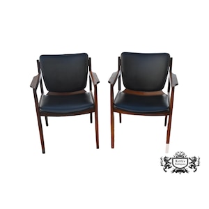 May include: Two mid-century modern armchairs. Each chair has a dark, possibly black, leather seat and backrest. The armrests and legs are made of wood, with a warm brown finish. The chairs are set against a white background.