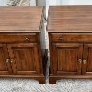May include: Two matching wooden nightstands with a rich, dark brown finish. Each nightstand features a drawer, two cabinet doors, and brass-toned hardware. The nightstands are placed side by side.
