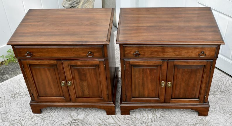 May include: Two matching wooden nightstands with a rich, dark brown finish. Each nightstand features a drawer, two cabinet doors, and brass-toned hardware. The nightstands are placed side by side.