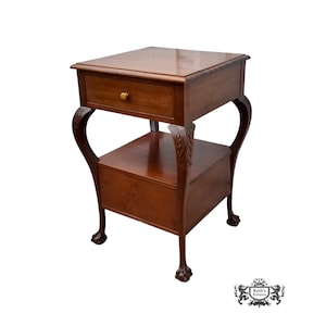 May include: A two-tiered, antique wooden side table with a drawer and a lower shelf. The table has curved legs and claw feet. The drawer has a brass knob. The wood has a rich, warm brown color.