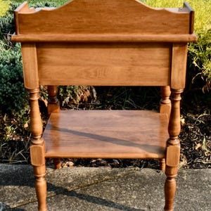 May include: A wooden side table with a warm, medium-brown finish. The table features a curved back, a lower shelf, and turned legs. The design is traditional, with a focus on the wood grain and craftsmanship. The table is suitable for a living room or bedroom.