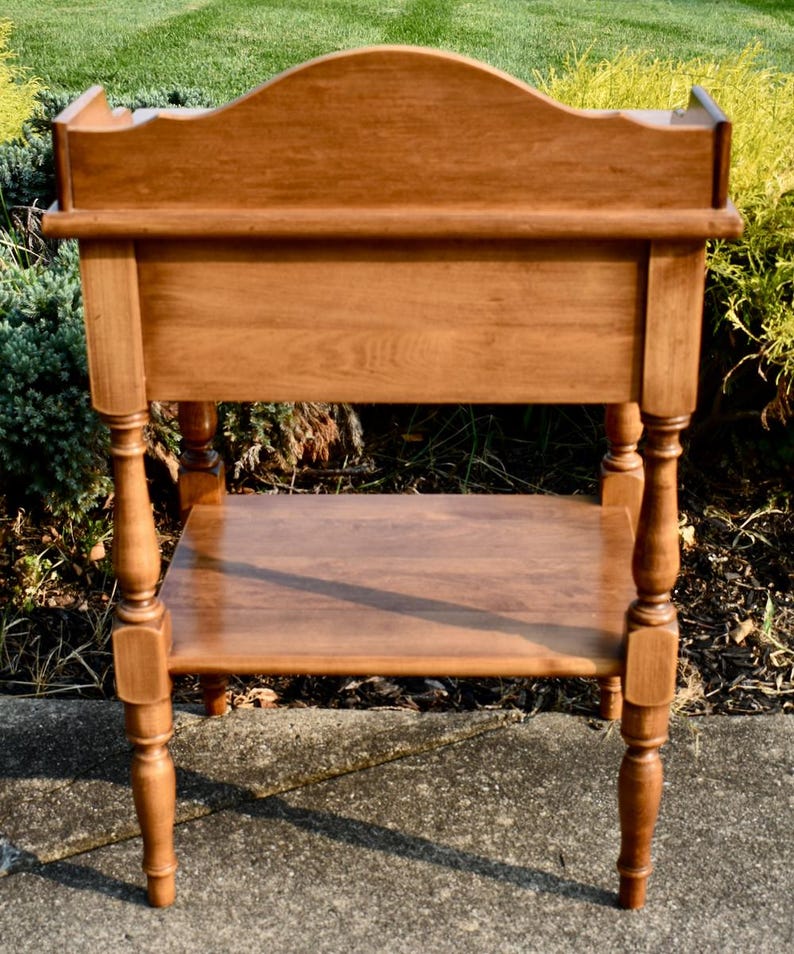 May include: A wooden side table with a warm, medium-brown finish. The table features a curved back, a lower shelf, and turned legs. The design is traditional, with a focus on the wood grain and craftsmanship. The table is suitable for a living room or bedroom.