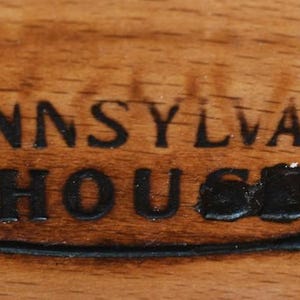 May include: A close-up of a wooden plaque with the words "PENNSYLVANIA HOUSE" in black, within an oval frame. The plaque is set against a wood grain background, showcasing the natural texture and color variations of the wood.