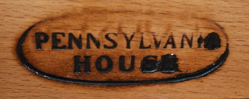 May include: A close-up of a wooden plaque with the words "PENNSYLVANIA HOUSE" in black, within an oval frame. The plaque is set against a wood grain background, showcasing the natural texture and color variations of the wood.