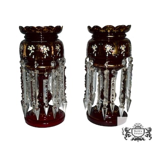 May include: Pair of antique ruby red glass candle holders with crystal prism accents. Each holder features a scalloped rim, floral and gold detailing, and a central column. The crystal prisms hang from the body of the holders.