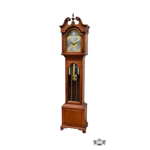 May include: A tall wooden grandfather clock with a swan neck pediment, a brass-toned clock face featuring Roman numerals, and black hands. The central glass panel reveals three hanging brass weights and a pendulum, set above a solid base.