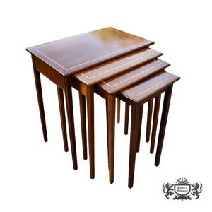 May include: A set of four nested wooden tables with a polished brown finish. Each rectangular tabletop features a light-colored inlaid border. The tables are arranged in a cascading formation, each supported by four straight, slender wooden legs.