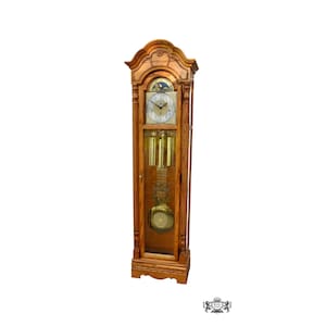 May include: A tall wooden grandfather clock with a light brown finish, featuring a glass-fronted door, a white clock face with black numerals, a moon phase dial, three hanging brass weights, and a large circular brass pendulum inside the case.