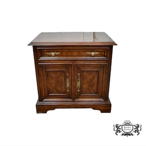 May include: A dark brown wooden nightstand with two doors and a drawer. The nightstand has brass hardware and a glossy finish. The doors and drawer feature decorative wood grain patterns. The nightstand is a classic piece of furniture.