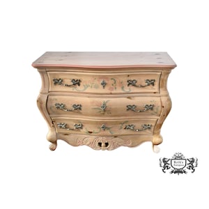 May include: A three-drawer antique chest of drawers with a light beige finish. The chest features curved sides, ornate metal handles, and floral painted accents in pink and green. The top has a pink trim.