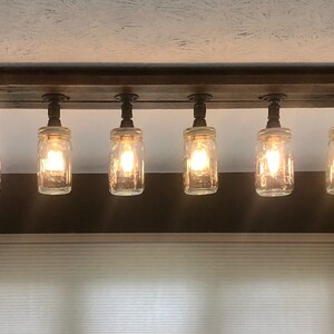 Pu&ograve; includere: Un lampadario rustico per soffitto con sei barattoli di vetro trasparente appesi a una trave di legno. I barattoli hanno lampadine all'interno e sono cablati con fili elettrici neri.