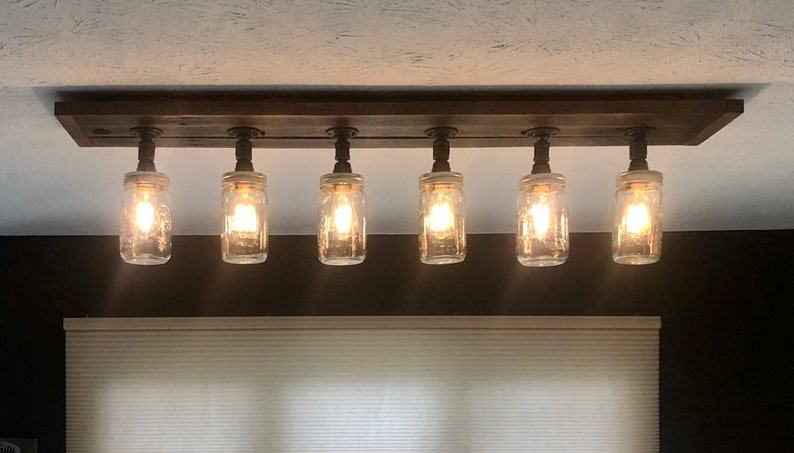 Pu&ograve; includere: Un lampadario rustico per soffitto con sei barattoli di vetro trasparente appesi a una trave di legno. I barattoli hanno lampadine all'interno e sono cablati con fili elettrici neri.