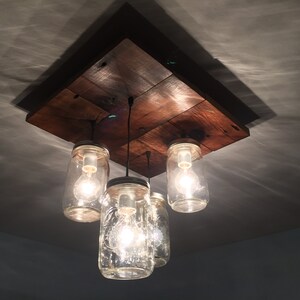 May include: A rustic wooden ceiling light fixture with four glass mason jars used as light shades. The jars have clear glass and contain Edison light bulbs.