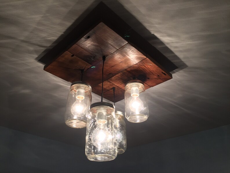 May include: A rustic wooden ceiling light fixture with four glass mason jars used as light shades. The jars have clear glass and contain Edison light bulbs.