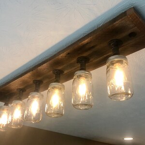 Pu&ograve; includere: Un apparecchio di illuminazione rustico in stile farmhouse con sei barattoli di vetro trasparente appesi a una tavola di legno con tubi di metallo nero. I barattoli hanno lampadine all'interno e sono accesi.