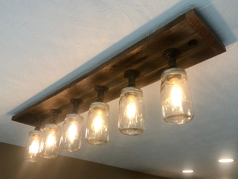 Pu&ograve; includere: Un apparecchio di illuminazione rustico in stile farmhouse con sei barattoli di vetro trasparente appesi a una tavola di legno con tubi di metallo nero. I barattoli hanno lampadine all'interno e sono accesi.