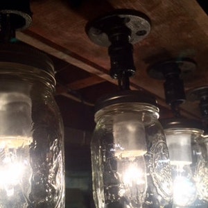 Pu&ograve; includere: Primo piano di un lampadario fatto di barattoli di vetro con lampadine a vista. I barattoli sono sospesi da una trave di legno con tubi di metallo nero.