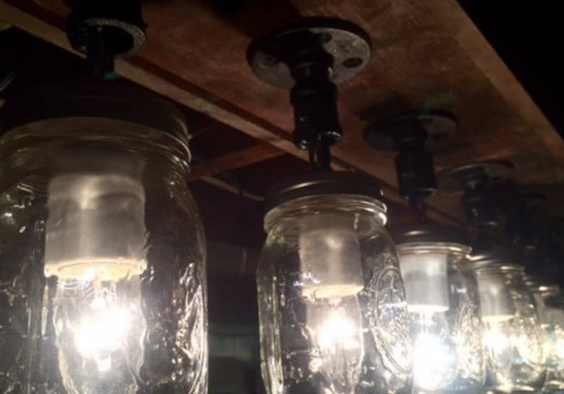 Pu&ograve; includere: Primo piano di un lampadario fatto di barattoli di vetro con lampadine a vista. I barattoli sono sospesi da una trave di legno con tubi di metallo nero.