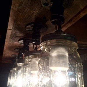 Pu&ograve; includere: Un apparecchio di illuminazione rustico in stile farmhouse realizzato con barattoli di vetro e raccordi in tubo nero. I barattoli sono appesi al soffitto con catene e contengono lampadine.