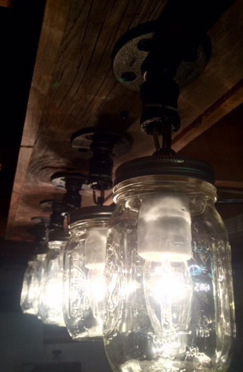 Pu&ograve; includere: Un apparecchio di illuminazione rustico in stile farmhouse realizzato con barattoli di vetro e raccordi in tubo nero. I barattoli sono appesi al soffitto con catene e contengono lampadine.