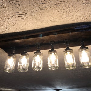 Pu&ograve; includere: Un lampadario rustico in legno per soffitto con sei barattoli di vetro trasparente e lampadine Edison. Il lampadario &egrave; montato su una trave di legno.