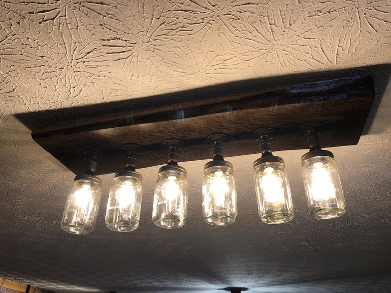 Pu&ograve; includere: Un lampadario rustico in legno per soffitto con sei barattoli di vetro trasparente e lampadine Edison. Il lampadario &egrave; montato su una trave di legno.